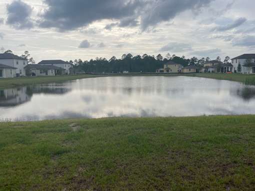 A serene landscape with a lake, houses, and trees under a cloudy sky.