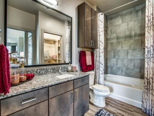 	Grand Prairie, TX Apartments - Waters Edge at Mansfield - Bathroom With Wood-Style Flooring, Ceramic Tub, Framed Vanity Mirrors With Decorative Lighting, and Granite Countertop.