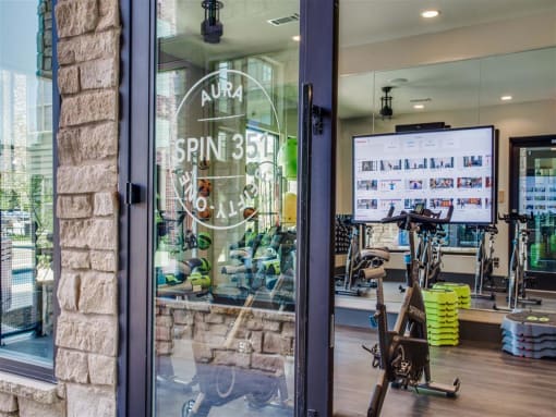 Apartments In Grand Prairie, TX - Waters Edge At Mansfield - Exterior View Of Spin Area With Wood Style Flooring, Large Windows, And TV