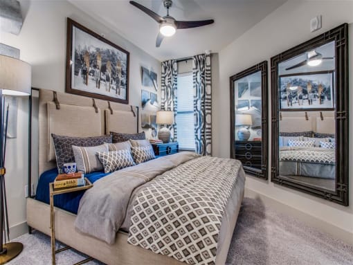 	Dog Friendly Apartments in Grand Prairie, TX - Waters Edge at Mansfield - Bedroom With Custom Carpeting, Ceiling Fan, Window With 2" Blinds, Floor Lamp, Wall Art, Wall Mirrors, and Nightstand With La