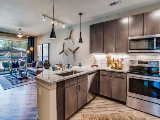 Pet Friendly Apartments In Grand Prairie, TX - Waters Edge At Mansfield - Modern Kitchen With Gray Wood Cabinets, Modern Decor, Wood-Style Flooring, Granite Countertops, And Stainless Steel Appliances