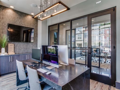 	Pet Friendly Apartments in Grand Prairie, TX - Waters Edge at Mansfield - Private Business Center With Table, Chairs, Computers, Mounted TV, Pendant Light Fixture, and Puck Lighting.