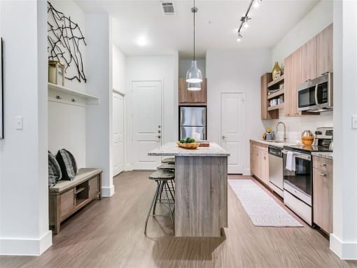 Apartments for Rent in Rowlett, TX - Village of Rowlett - Kitchen with Wood-Style Flooring, Granite Countertops, Wood-Tone Cabinetry, Stainless Steel Appliances, Kitchen Island/Breakfast Bar, Bar-Style Stools, Pendant Lights, Pantry, Storage Shelves, and Bench Seat.