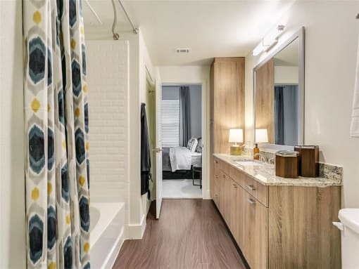 Rowlett, TX Apartments for Rent - Village of Rowlett - Bathroom With Wood-Style Flooring, Shower and Tub Combo, Large Vanity Mirror, Wide Granite Countertop with Sink and Wood-Style Cabinets
