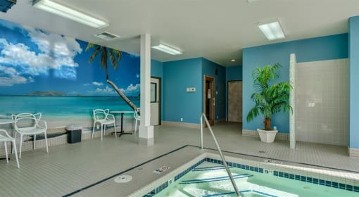 Apartments for Rent in Spokane, WA - Canyon Bluffs - Indoor Pool with Tiled Flooring, White Table with Chairs, Potted Plant, and Wall Mural of Beach Scenery.