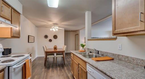 Spokane, WA, Apartments - Canyon Bluffs - Fully Equipped Kitchen with Appliances, Granite Countertops, Wooden Cabinetry, Wood-Style Flooring, Dual Sink, and View of Dining Area with a Table and Chairs.