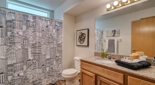 Apartments for Rent Spokane, WA - Canyon Bluffs - Bathroom with Wood-Style Flooring, Shower Curtain, Window, Vanity with Sink, Granite Countertops, Wood-Style Cabinetry, and Large Mirror.