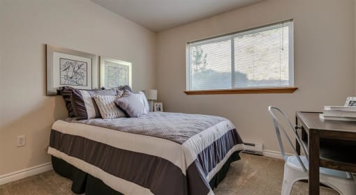 Apartments in Spokane, WA - Canyon Bluffs - Bedroom with Carpeted Flooring, Large Bed, Nightstand with Lamp, Wooden Desk, White Chair, Beige Walls, and Large Window.