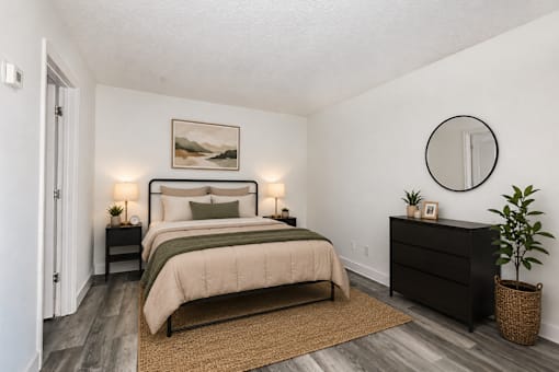 A bedroom with a large bed, a nightstand, a lamp, a plant, and a round mirror on the wall.
