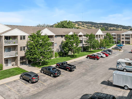 A parking lot with cars and a trailer in front of apartment buildings.