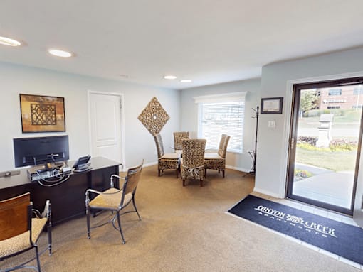 A living room with a black desk and chairs, a television, and a rug that says