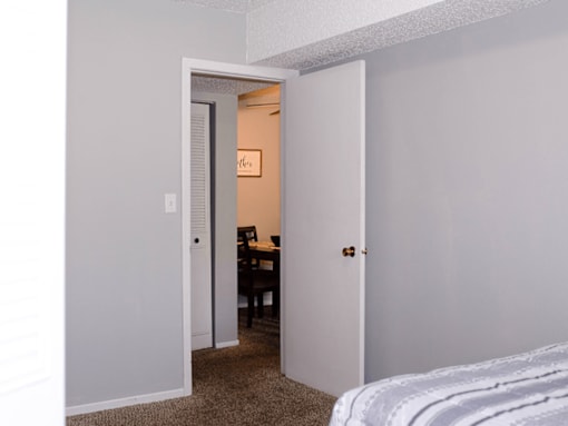 A white door opens to a room with a desk and a bed.