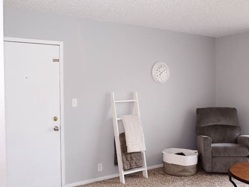 A white door is on the left of a room with a ladder, a chair, and a clock on the wall.