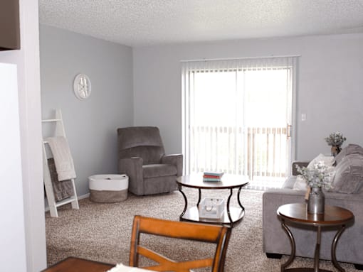 A living room with a grey couch, a brown chair, a grey armchair, a white ottoman, a small table, and a window with white curtains.