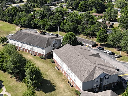 A large white building with a black roof is surrounded by trees and smaller buildings.