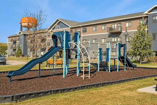 A playground with a blue slide and a brown sandbox in front of a building.