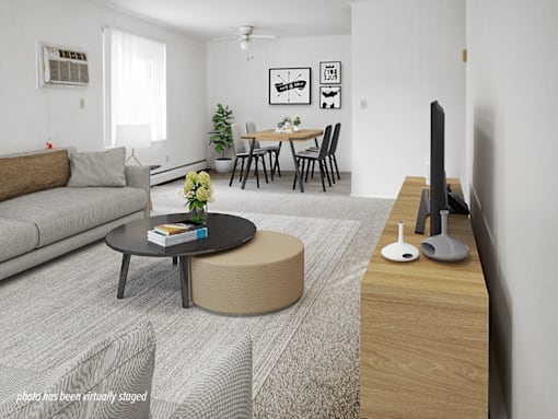A living room with a grey sofa, a black coffee table, a round ottoman, a wooden TV stand, and a dining table with chairs.
