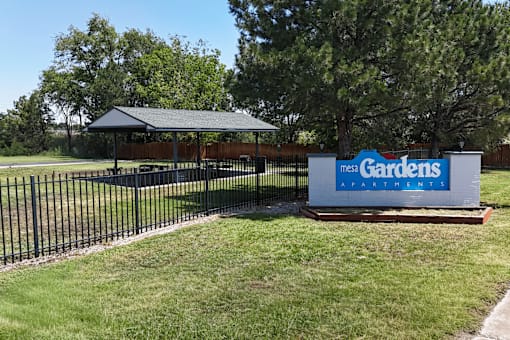A sign for Mega Gardens stands in front of a fence and trees.