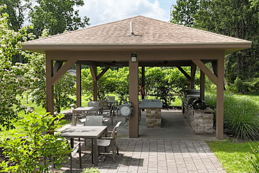 A wooden gazebo with a table and chairs is surrounded by greenery.