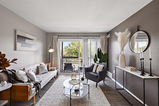 A living room with a grey couch, a glass coffee table, a grey armchair, a grey side table, a grey cabinet, a round mirror, a framed picture, a potted plant, a window with curtains,