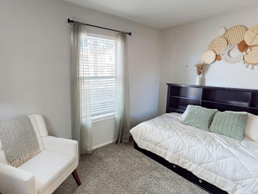 A white chair sits in a bedroom with a bed and a window with curtains.