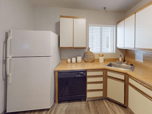 A white refrigerator stands in a kitchen with wooden cabinets and a black dishwasher.