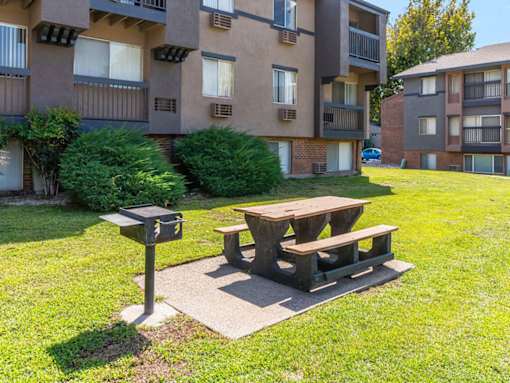 A wooden picnic table sits in the grass in front of apartment buildings.