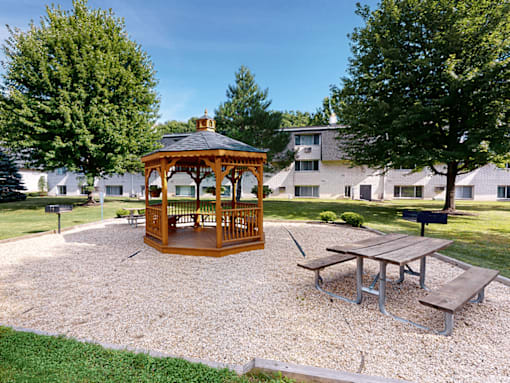 A gazebo is in the middle of a gravel area with a picnic table and benches.