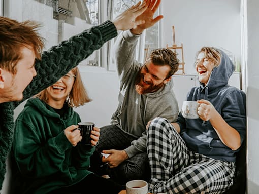 Four people in pajamas are sitting on a couch, laughing and holding mugs.