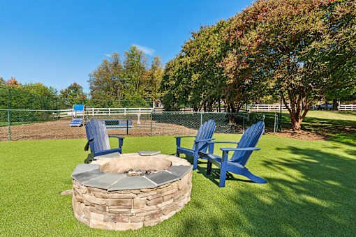 A stone fire pit sits in the middle of a grassy area with blue chairs around it.