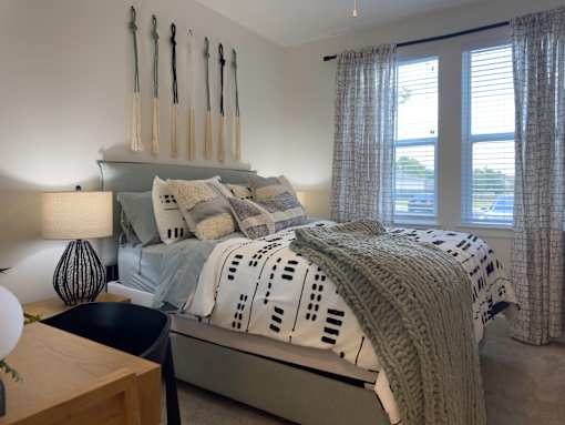 Luxury Apartments in Myrtle Beach, SC, With In-Unit Washer and Dryer - Anchor Pointe - Cozy Bedroom With Upholstered Bed, Patterned Bedding, Bedside Lamp, Wall Decor, and Windows With Curtains.