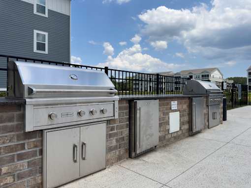 Dog-Friendly Luxury Apartments in Myrtle Beach, SC - Anchor Pointe - Outdoor Grilling Station With Stainless Steel Gas Grills, Brick Counter, Fenced Patio Pathway, and Overlooking Apartment Homes.