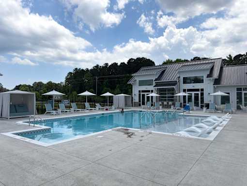 2-Bedroom Luxury Apartments in Myrtle Beach, SC - Anchor Pointe - Resort-Style Swimming Pool With In-Water Loungers, Sun Deck Chairs, Umbrella Seating, White Cabanas, and Clubhouse Building.