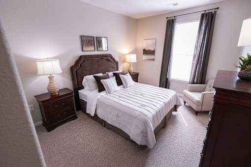 A bedroom with a large bed, a nightstand with a lamp, a chair, and a window with curtains.