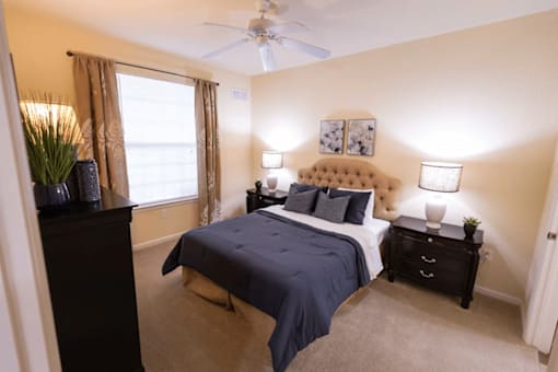Dog-Friendly Apartments in Asheville, NC - Reserve at Asheville - Bedroom with Carpet Flooring, Bed, Nightstands, Table Lamps, Dresser, Wall Arts, Ceiling Fan, and Large Window with Coverings.