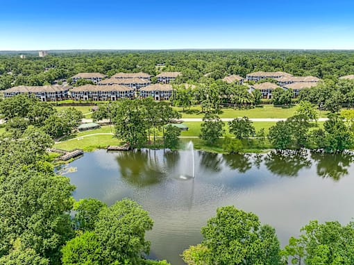 A serene landscape with a fountain in the middle of a lake surrounded by lush green trees and buildings in the distance.
