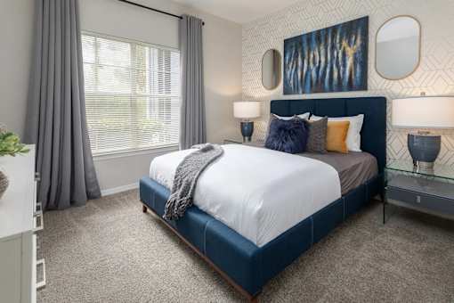 A bedroom with a large bed, pillows, nightstands with lamps, and a large window with grey curtains hanging.