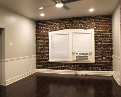 Living room in Brookmore apartments Pasadena CA. Large window with white blinds and trim and air conditioning unit on an exposed brick wall