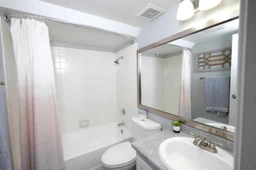 Fusion Fort Worth studio apartments Fort Worth. Bathroom with white tile tub and ombre shower curtain, white toilet, and single sink with vanity light and mirror.
