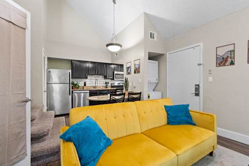 A yellow couch with a blue pillow is in the foreground of a kitchen and living room at Fusion Orlando furnished apartments.