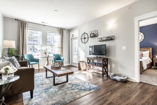 A furnished living room at The Fort with a TV, stylish decor, hardwood-style flooring, and windows, next to a bedroom.