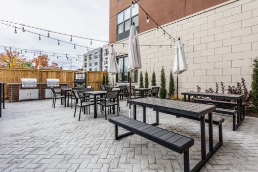 One BR Apartments in Cincinnati, OH - Madamore - Grill Area with BBQs, Tables, Chairs, Umbrellas, and String Lights.