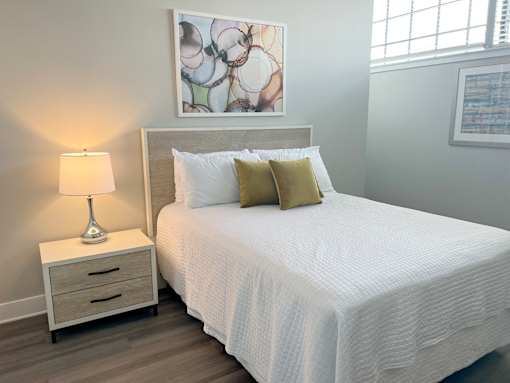 A bed with a white comforter and two pillows sits next to a nightstand with a lamp on it.