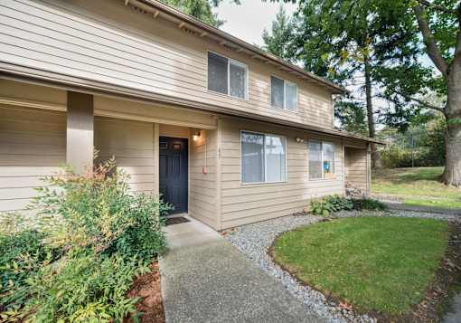 A house with a brown siding and a black door is surrounded by greenery.