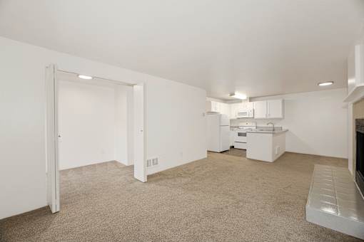 A white room with a carpet floor and a kitchen area in the background.