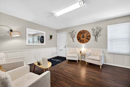 A living room with a grey couch, a white armchair, a wooden coffee table, and a large clock on the wall.