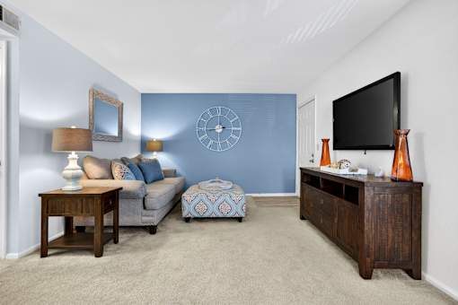 A living room with a grey couch, a wooden table, a blue ottoman, a clock, a flat screen TV, and a vase.