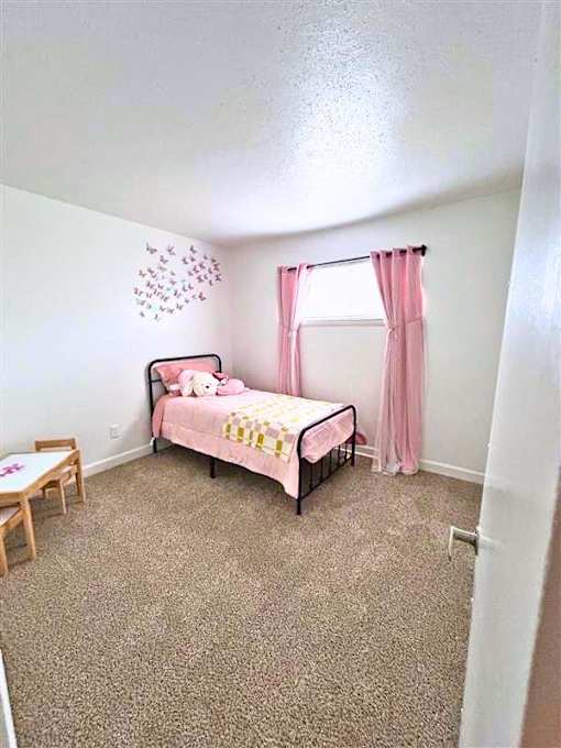 A pink bed with a teddy bear on it sits in a room with a carpeted floor.