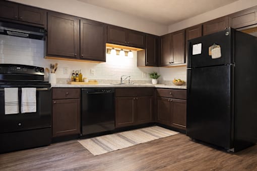 A black refrigerator stands in a kitchen with brown cabinets and a white backsplash.