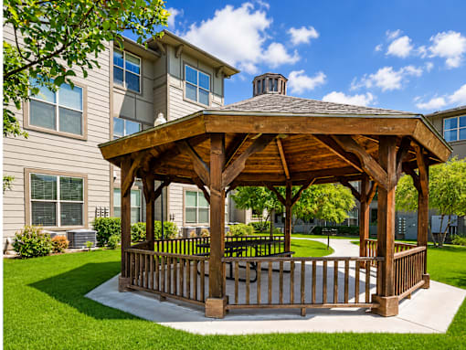 A wooden gazebo is situated in a grassy area in front of a building.